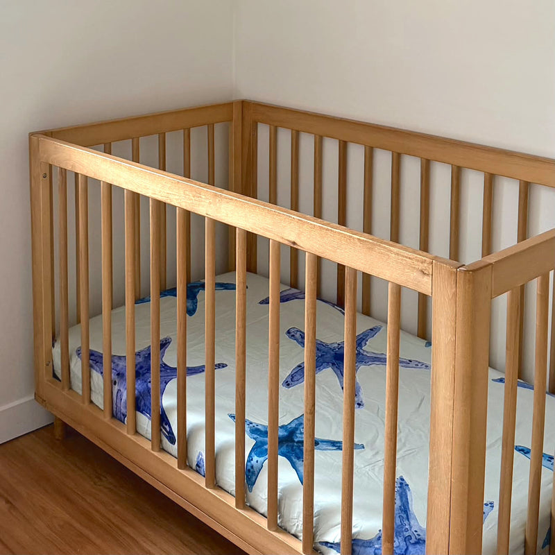 A light natural wood crib with vertical slats, styled with a white mattress and blue starfish-patterned bedding, set against a white wall with a small framed print on the corner.