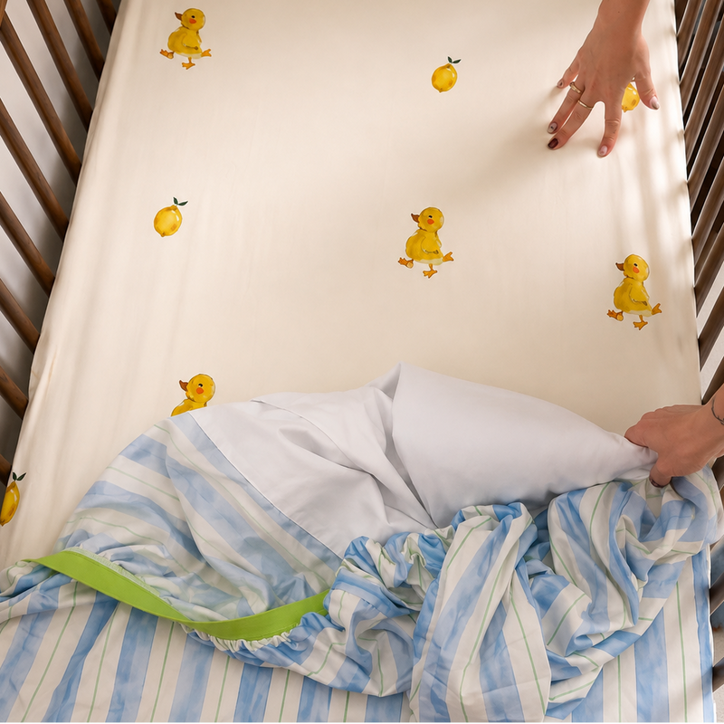 A baby crib mattress fitted with an off-white sheet printed with small yellow ducklings, shown in a wooden crib with a person adjusting a blue and white striped blanket.